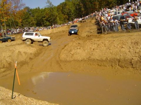 1979 Ford Bronco - Off-road vehicles navigate a muddy track during an event, with a crowd watching from the hillside.