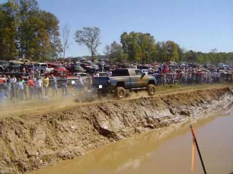 1979 Ford Bronco - A muddy pickup truck navigates a challenging mud pit during an event, with a large crowd watching from the sidelines.