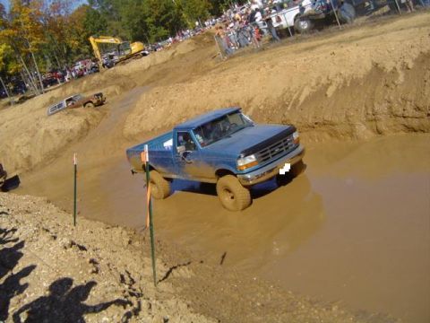 1979 Ford Bronco - A blue pickup truck tackles a muddy obstacle course during an event with onlookers present.