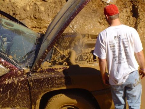 1979 Ford Bronco - A maroon SUV is deeply stuck in mud, its hood propped open to reveal the engine. A man in a red cap and white t-shirt stands nearby, observ…