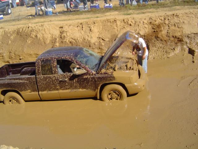 Mud-covered pickup truck stuck in a mud pit with hood open, person looking at engine