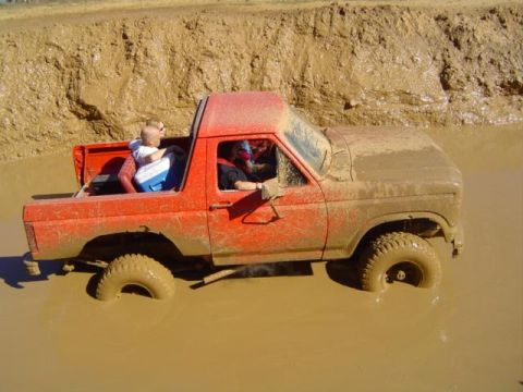 1979 Ford Bronco - A red SUV is stuck in a mud pit with two people inside.