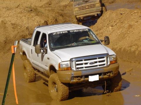 1979 Ford Bronco - A white pickup truck navigates a muddy obstacle course, with another vehicle visible in the background.
