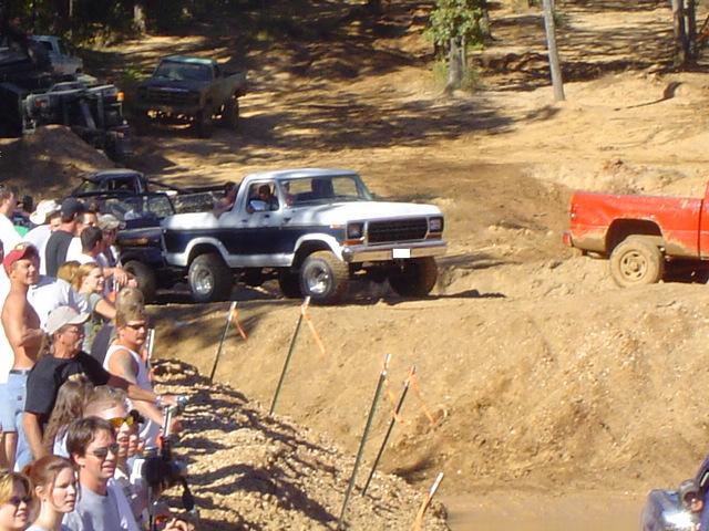 White and blue Ford Bronco driving on a dirt track with a crowd watching. A red truck is to the right