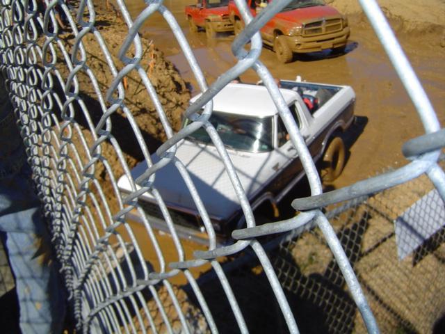 White and brown pickup truck stuck in mud, viewed through a chain-link fence