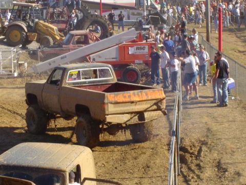 1979 Ford Bronco - A muddy pickup truck is being pulled by a chain in a dirt arena, with spectators watching from behind a fence.