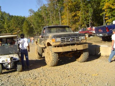 1979 Ford Bronco - A heavily muddied pickup truck sits on a dirt track, with other vehicles and trees in the background.