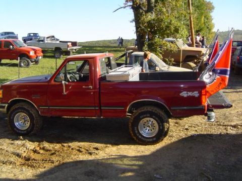 1979 Ford Bronco - A red pickup truck is parked in a muddy area with several other vehicles visible in the background.