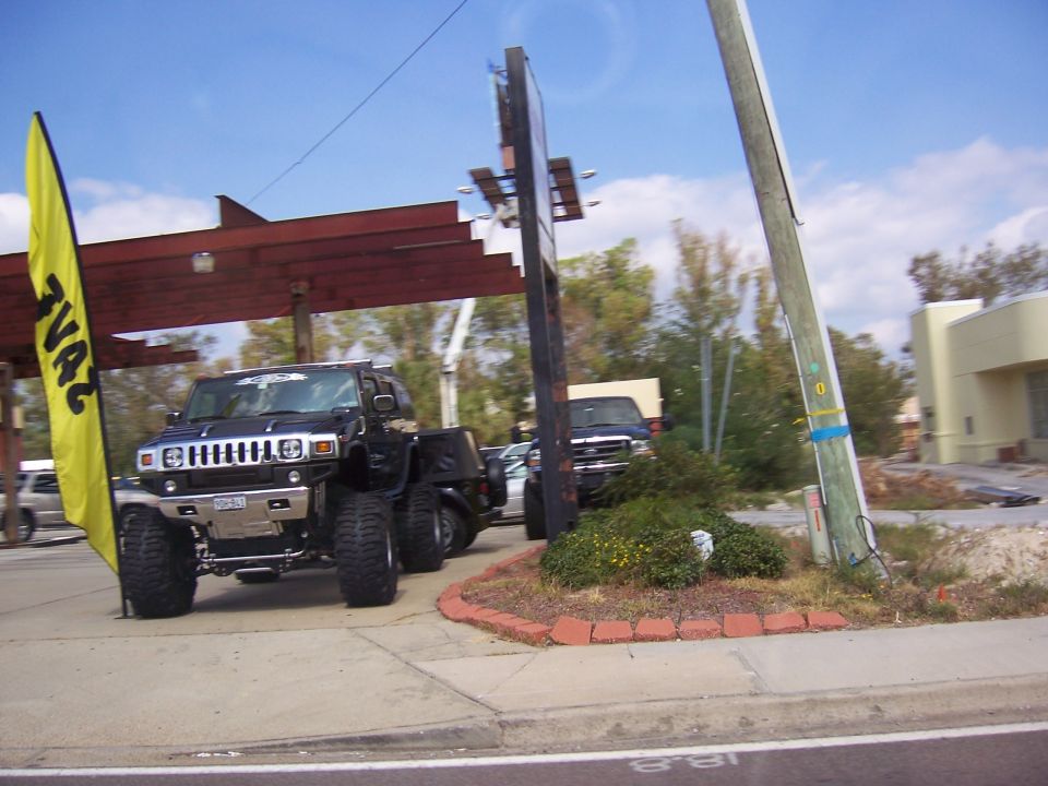 Black lifted six-wheel Hummer H2 conversion on big mud tires parked at a roadside commercial lot