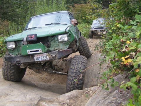 1990 Ford Bronco - A heavily modified green off-road vehicle tackles a challenging rocky terrain, with another vehicle visible in the background.