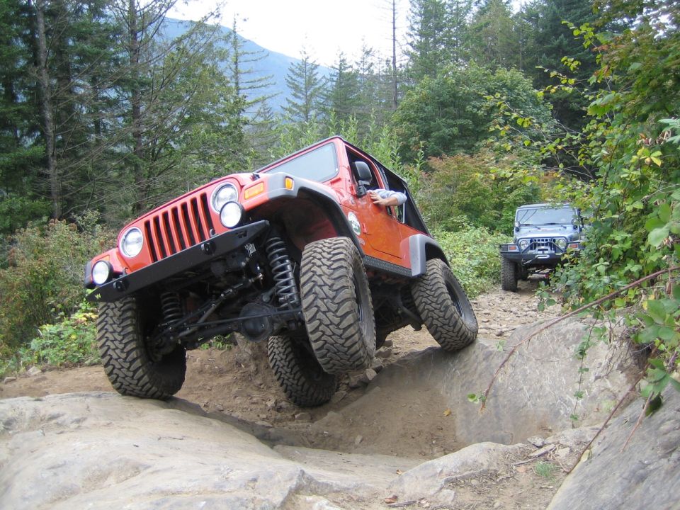 Orange Jeep Wrangler with lifted suspension and large tires navigating a rocky trail, with a second Jeep visible behind