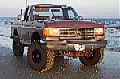 1987 Ford Bronco - Small low-resolution thumbnail of a lifted tan/brown Ford pickup with a brush guard and tall mud tires, photographed head-on at a muddy off…
