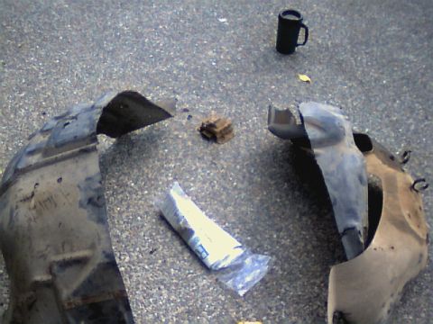 1987 Ford Bronco II - Two detached fender liners lie on the ground next to a plastic bag and a mug.
