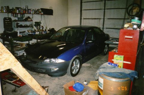 1996 Chevrolet Cavalier - A dark blue sedan with a black hood is parked in a cluttered garage.