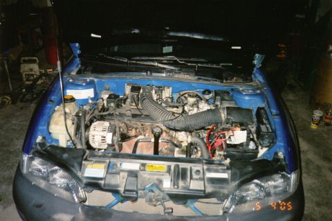 1996 Chevrolet Cavalier - Front-on view into the engine bay of a blue compact car in a garage with the hood up and the bumper cover and headlight trim removed. The t…