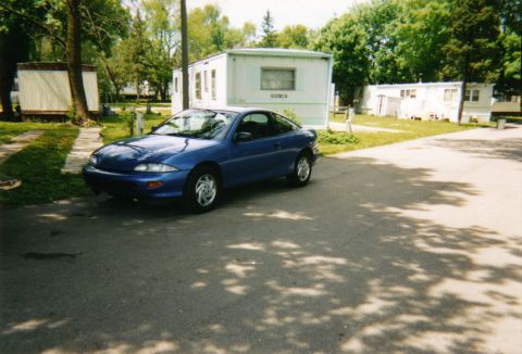 1996 Chevrolet Cavalier - A blue coupe sits parked on an asphalt road in front of several trailer homes on a sunny day.