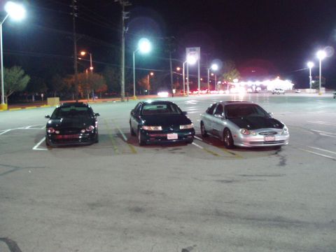 1993 Ford Taurus - Three sedans are parked in a lot at night under streetlights.