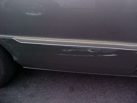 2000 Mercury Grand Marquis - Side view of a dark sedan showing damage to the lower door panel and chrome trim. The vehicle appears to be a 2000 Mercury Grand Marquis.