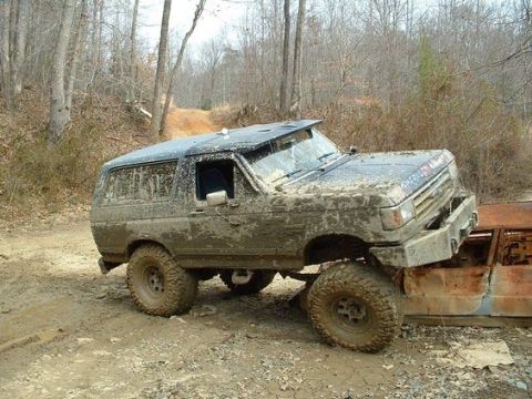 1988 Ford Bronco - This heavily muddied SUV, likely a 1987 model, is positioned on top of a rusted vehicle, suggesting off-road activity.