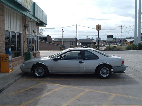1994 Ford Thunderbird - A silver 1994 Ford Thunderbird is parked in a lot next to a building with a newspaper stand.