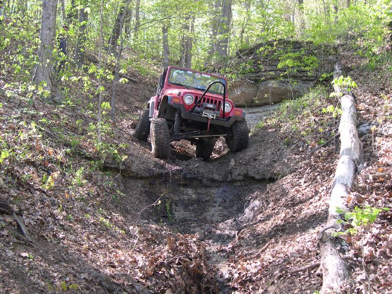 Red 1999 Jeep TJ navigating a steep, muddy off-road trail with trees on either side