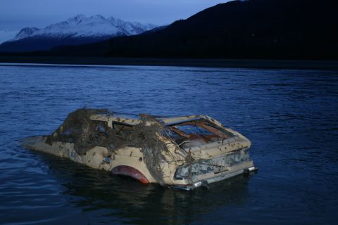 2003 Jeep TJ - A derelict vehicle sits partially submerged in a body of water, with debris clinging to its frame and mountains visible in the distance.
