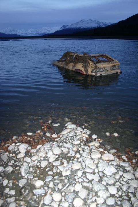 2003 Jeep TJ - A car sits partially submerged in a body of water, with a rocky shoreline in the foreground and mountains in the distance.