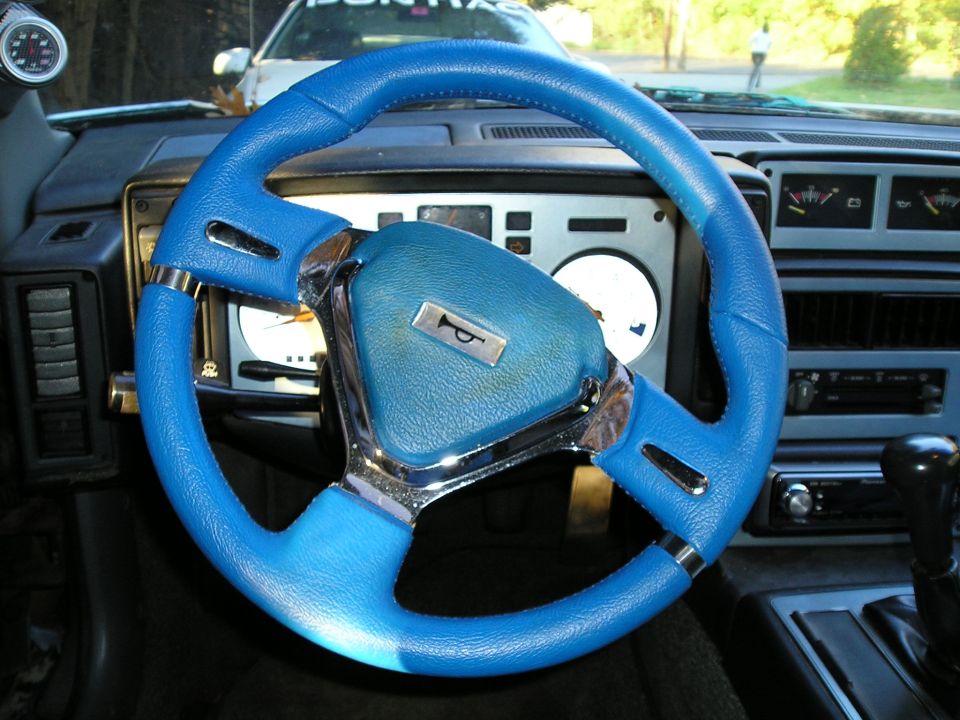 Interior shot of a car with a bright blue steering wheel and dashboard gauges. Another car is visible through the windshield