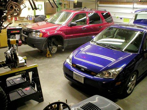 2003 Ford Escape - Garage interior with a red Ford Escape SUV up on a jack stand, front wheel removed and brake rotor exposed, parked beside a blue Ford Focus…
