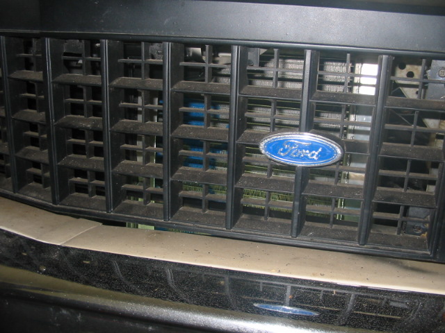 Close-up of a black grille with a blue Ford oval emblem. The grille has horizontal slats and a mesh behind it