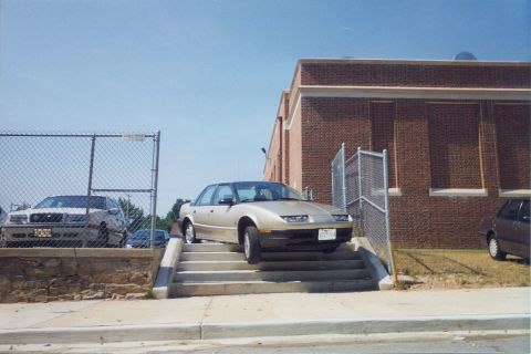 1998 Ford Crown Victoria - A gold sedan is precariously positioned on a set of outdoor stairs next to a brick building.