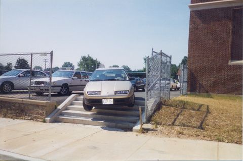 1998 Ford Crown Victoria - A gold Saturn SC sedan is precariously positioned on a set of stairs, with other vehicles visible in the background.