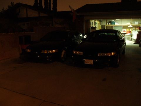 1995 Ford Taurus - Two dark-colored sedans are parked in front of a garage at night, with one car's license plate visible.