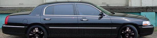 Side view of a black full-size sedan with tinted windows and black wheels parked by a weathered building