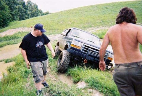 1995 Ford Bronco - A dark-colored 1995 Ford Bronco is stuck in a muddy off-road course, with two men observing the situation.