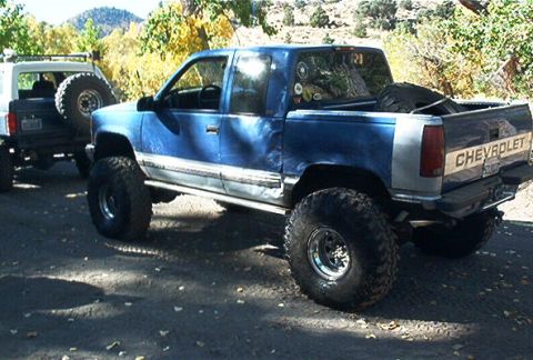 1986 Ford Bronco - Two lifted trucks, a blue Chevrolet and a white SUV, are parked on a dirt road with trees in the background.