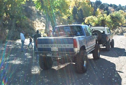 1986 Ford Bronco - Two off-road vehicles are parked on a dirt road with people gathered nearby on a sunny day.