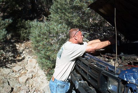 1986 Ford Bronco - A man works on the engine of a blue truck, its grille indicating an approximate 1980s model year.