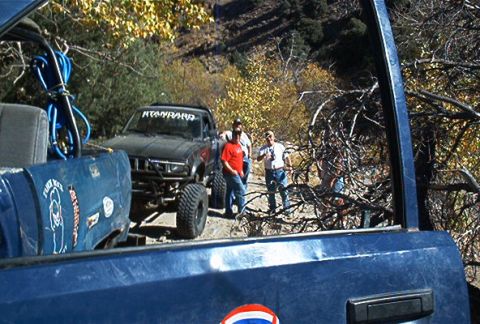 1986 Ford Bronco - Several people stand around a modified black pickup truck on a dirt road, surrounded by autumn foliage and dry brush.