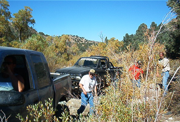Two pickup trucks parked in a dry, brushy area with people standing nearby. One truck has a banner on top