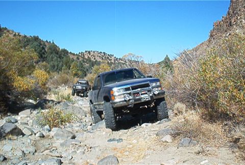 1986 Ford Bronco - Two trucks navigate a challenging, rocky path through a dry, brush-covered canyon.