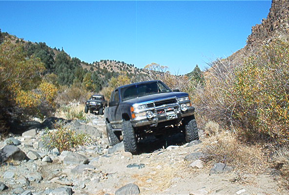 Blue pickup truck and another SUV driving on a rocky mountain trail under a clear blue sky