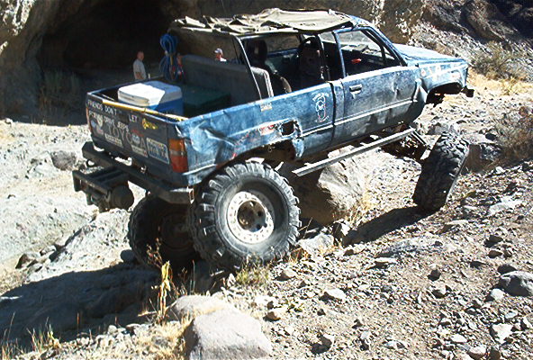 Weathered blue lifted mini-truck with cooler in bed climbing rocks in a desert wash