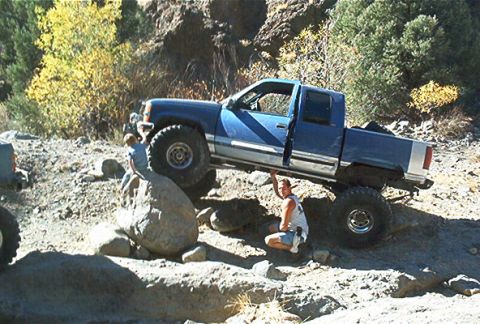 1986 Ford Bronco - A blue pickup truck navigates a challenging rocky off-road course with a person inspecting the undercarriage.