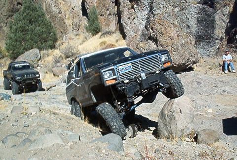 1986 Ford Bronco - A black Ford Bronco navigates a rocky terrain, with another vehicle and a person in the background.