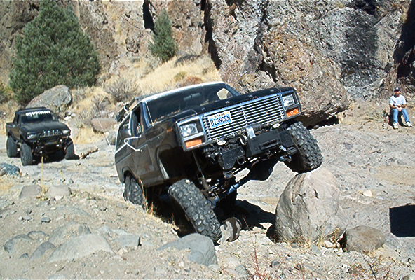 Black Ford Bronco off-roading over rocks, front grille visible, license plate reads BIGNRMY