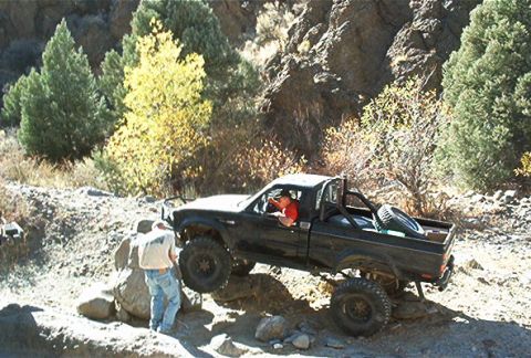 1986 Ford Bronco - Off-road pickup truck maneuvering over rocks with a person assisting.