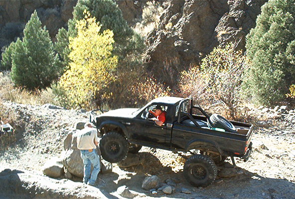 Black pickup truck with roll cage and spare tire in bed navigating rocky terrain, person standing by driver's side