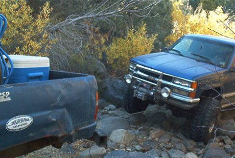 1986 Ford Bronco - A blue SUV is winched out of a rocky area by a dark blue pickup truck.