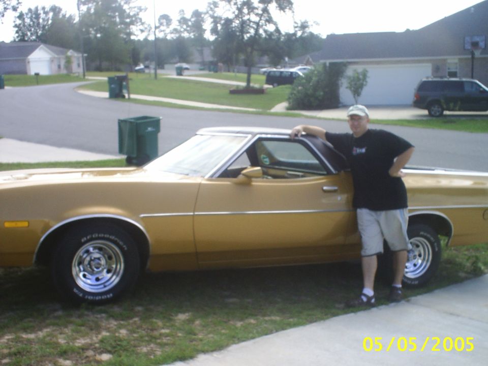 Gold Ford Ranchero with vinyl top and chrome slot wheels, owner leaning on open passenger door in a driveway